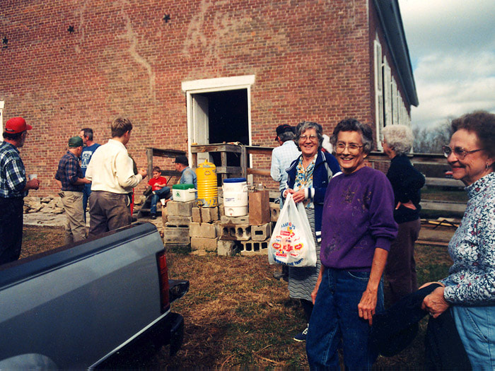 Women smile for the camera as men and women are outside the church where building supplies are stacked.