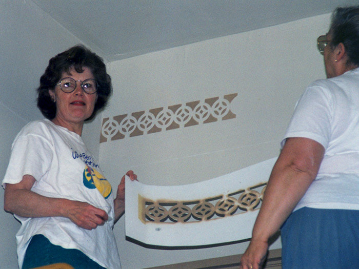 two women are painting the detail on the top of the wall with a template they are holding. One woman is looking into the camera contentedly